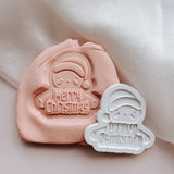 Gingerbread with santa hat holding merry Christmas debosser stamp and cutter